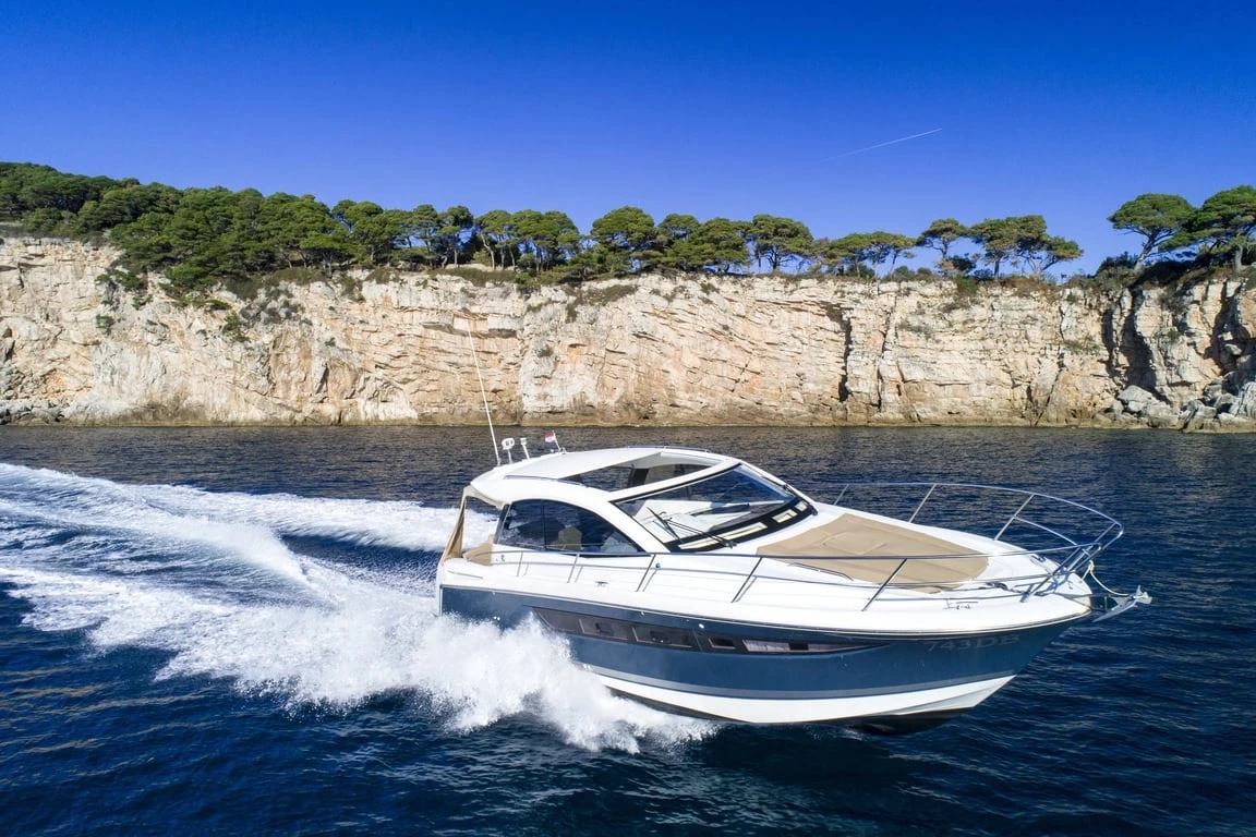 Jeanneau Leader 10 carving a turn with wake trailing, bow sun-pad visible, cliffs behind