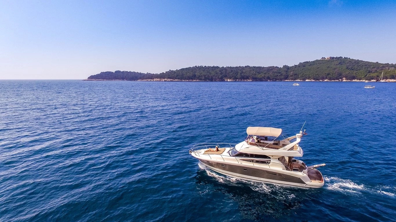 Prestige 440 Fly at slow cruise off a forested island, flybridge bimini up and teak swim platform visible astern